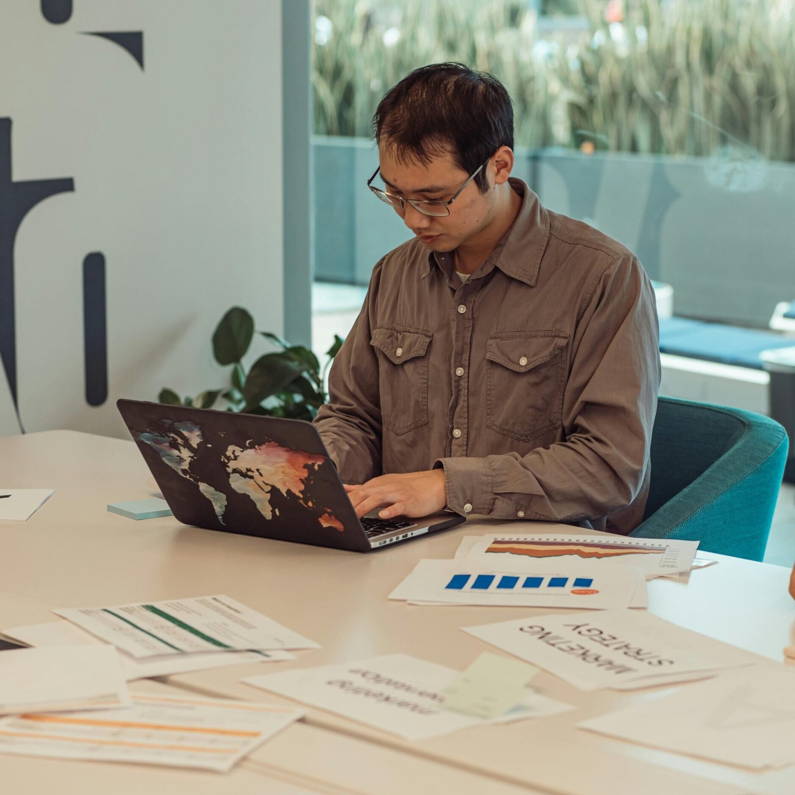 Focused Asian man working on a laptop surrounded by papers in a modern office setting.